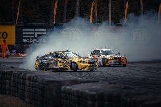  152 FIA drifta kauss, divas automašīnas dreifē pa trasi, no tām nāk dūmi, riepu, baneru ar tekstu, karogu un koku ieskautas fonā.